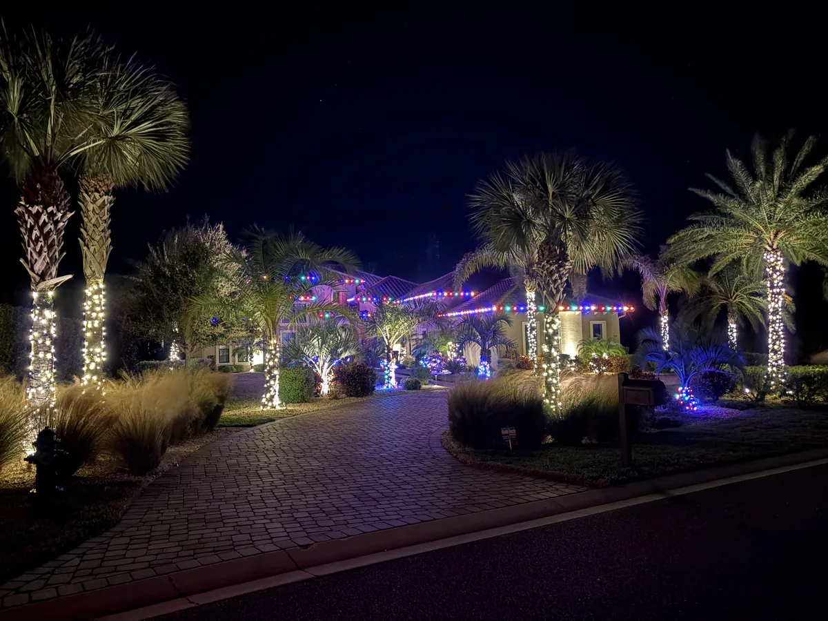 Large property with light-wrapped palms and a lit roofline along a paver driveway